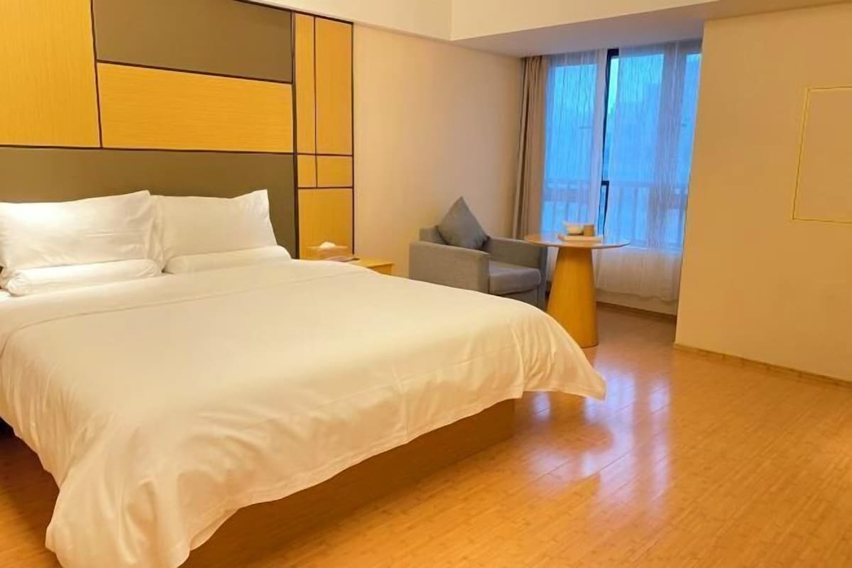 A hotel room with a large bed, a small table, a chair, and a window with curtains.