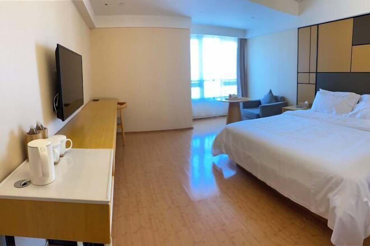 A hotel room with a bed, a TV, a desk with a kettle and mugs, a small table, and a window with a view.