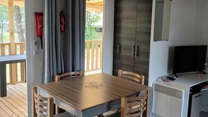 Een eetgedeelte met een houten tafel en stoelen, een kleine keuken met een televisie, en uitzicht op de omgeving door een schuifdeur.