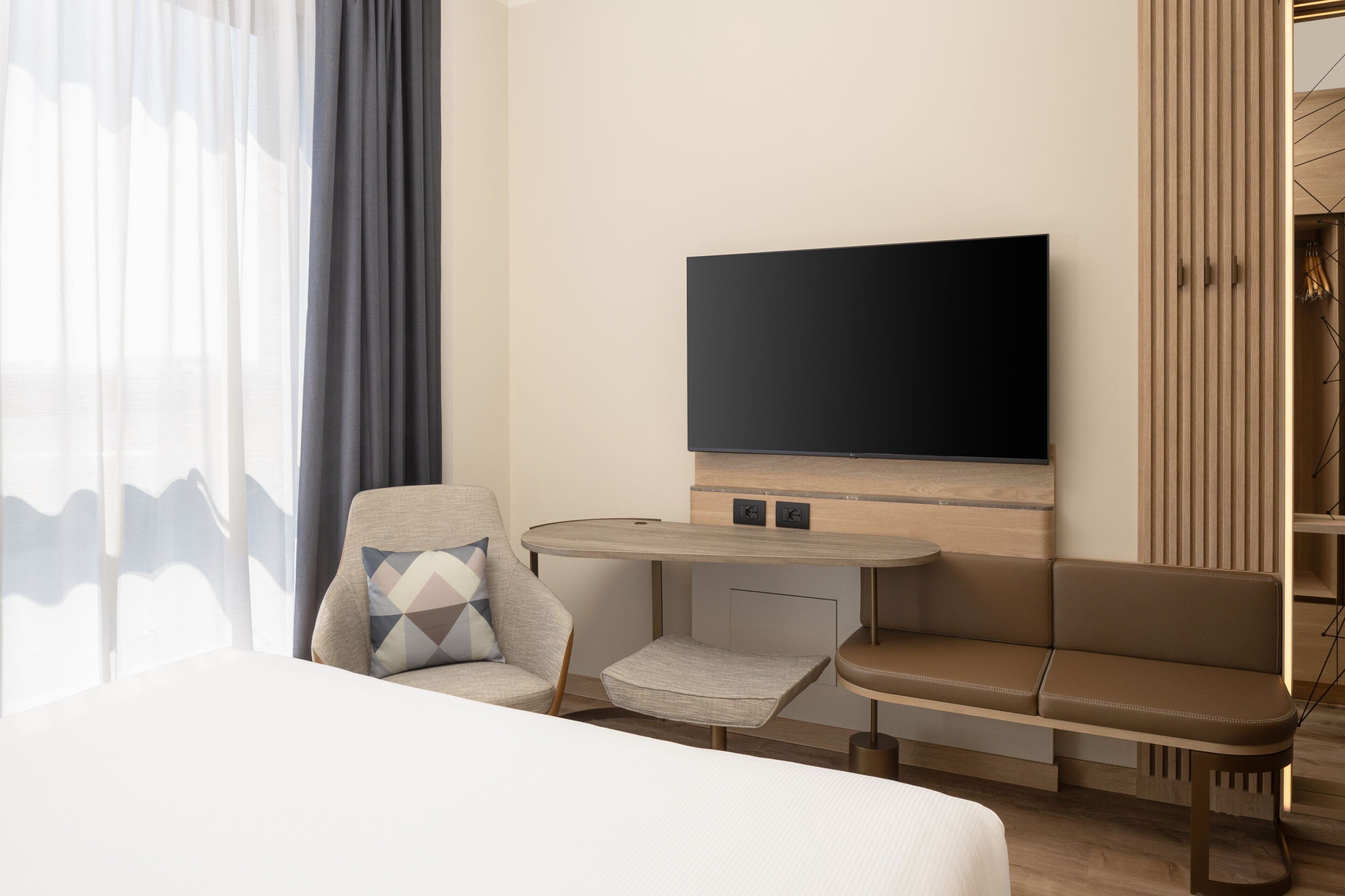 A modern hotel room with a large bed, a sofa, a small table, and a TV mounted on the wall.