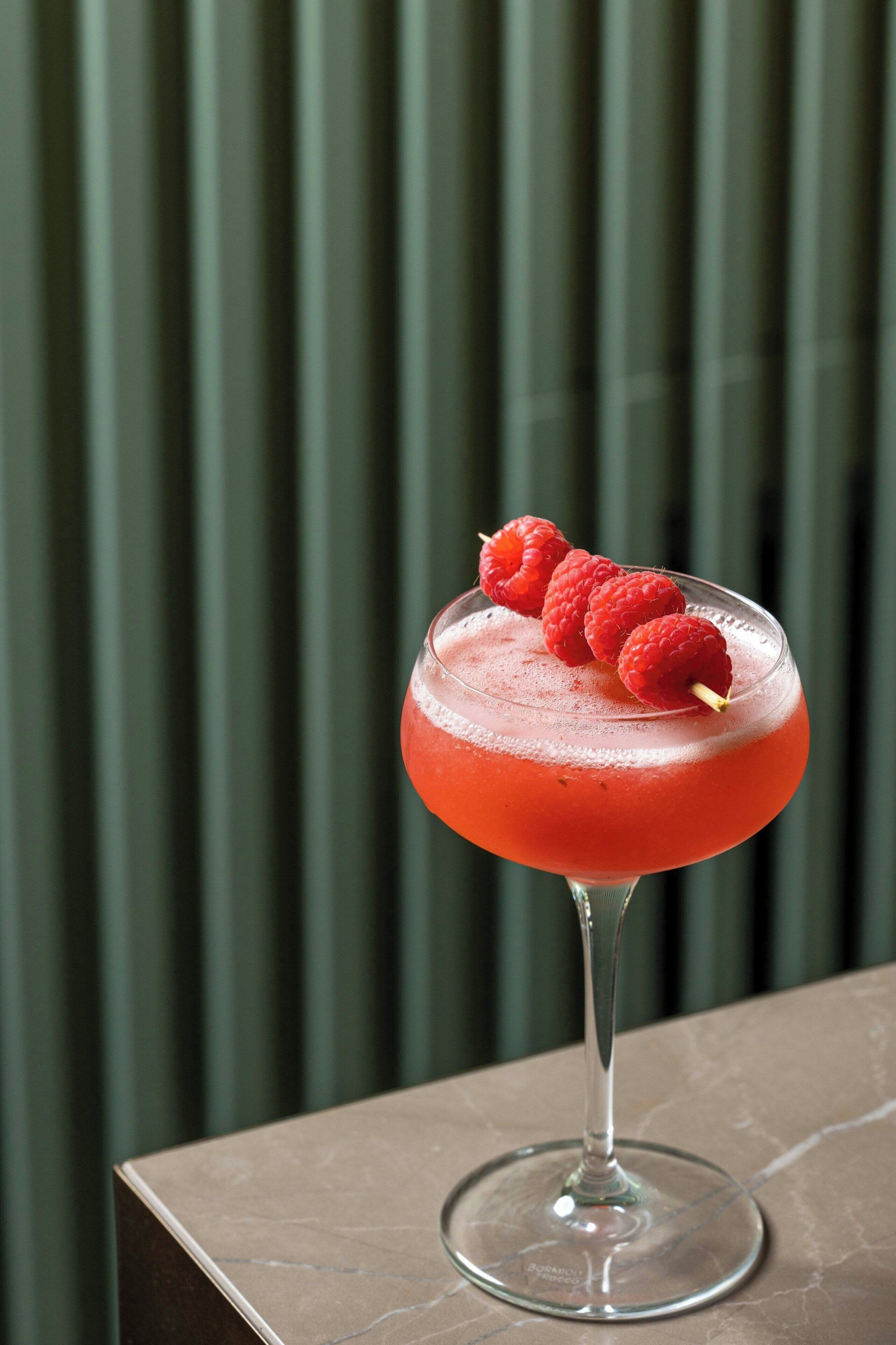 Un verre à cocktail contenant une boisson rouge et des framboises, posé sur une surface en marbre.