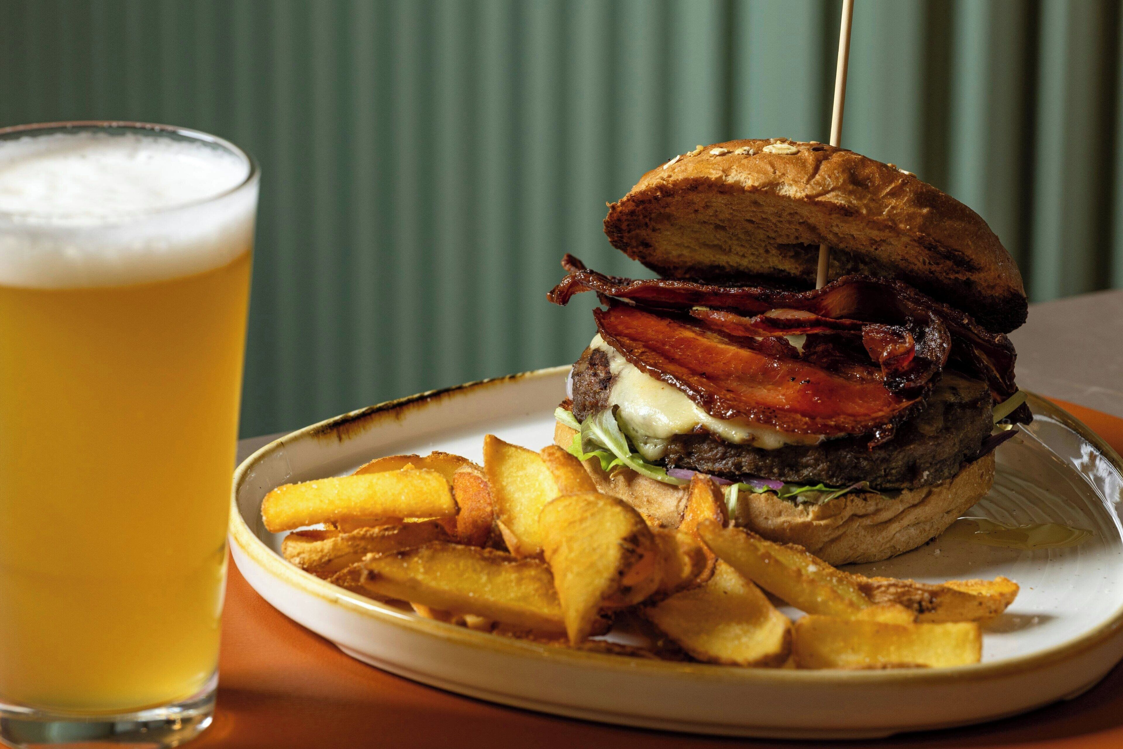 A burger with bacon, cheese, and lettuce on a bun, served with fries and a glass of beer.