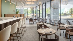 A modern restaurant interior with a bar area, seating arrangements, and large windows offering a view of the outdoors. - Courtyard By Marriott Milano Linate (Milan)