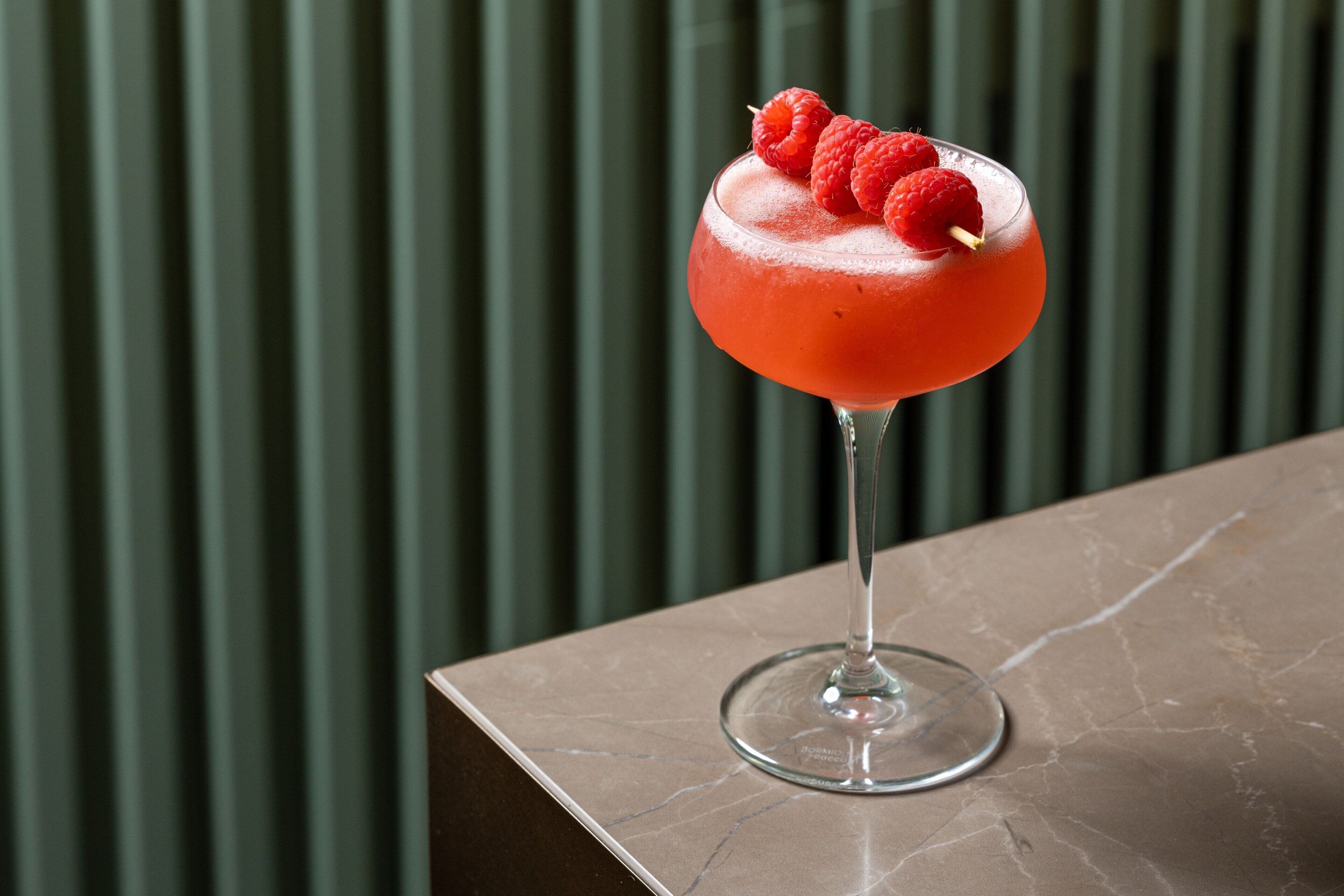 A cocktail in a glass with raspberries on top, placed on a marble surface.