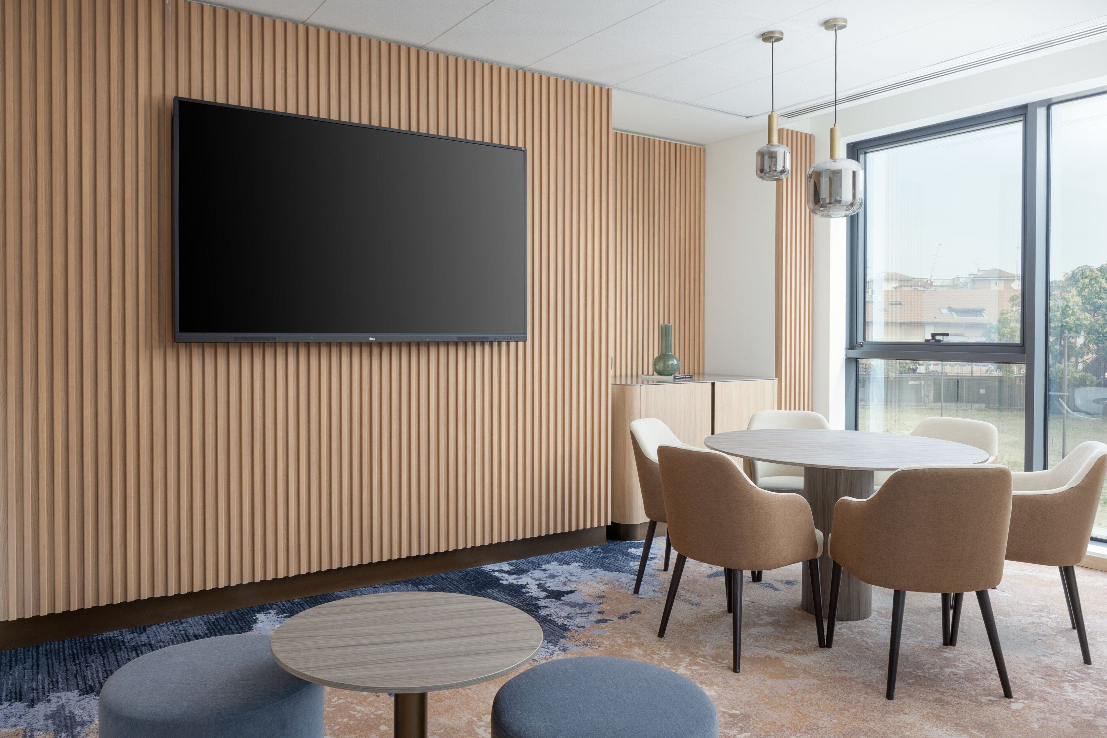 A modern conference room with a large flat-screen TV mounted on a wooden panel wall, a round table with six chairs, and a large window offering a view of the city.