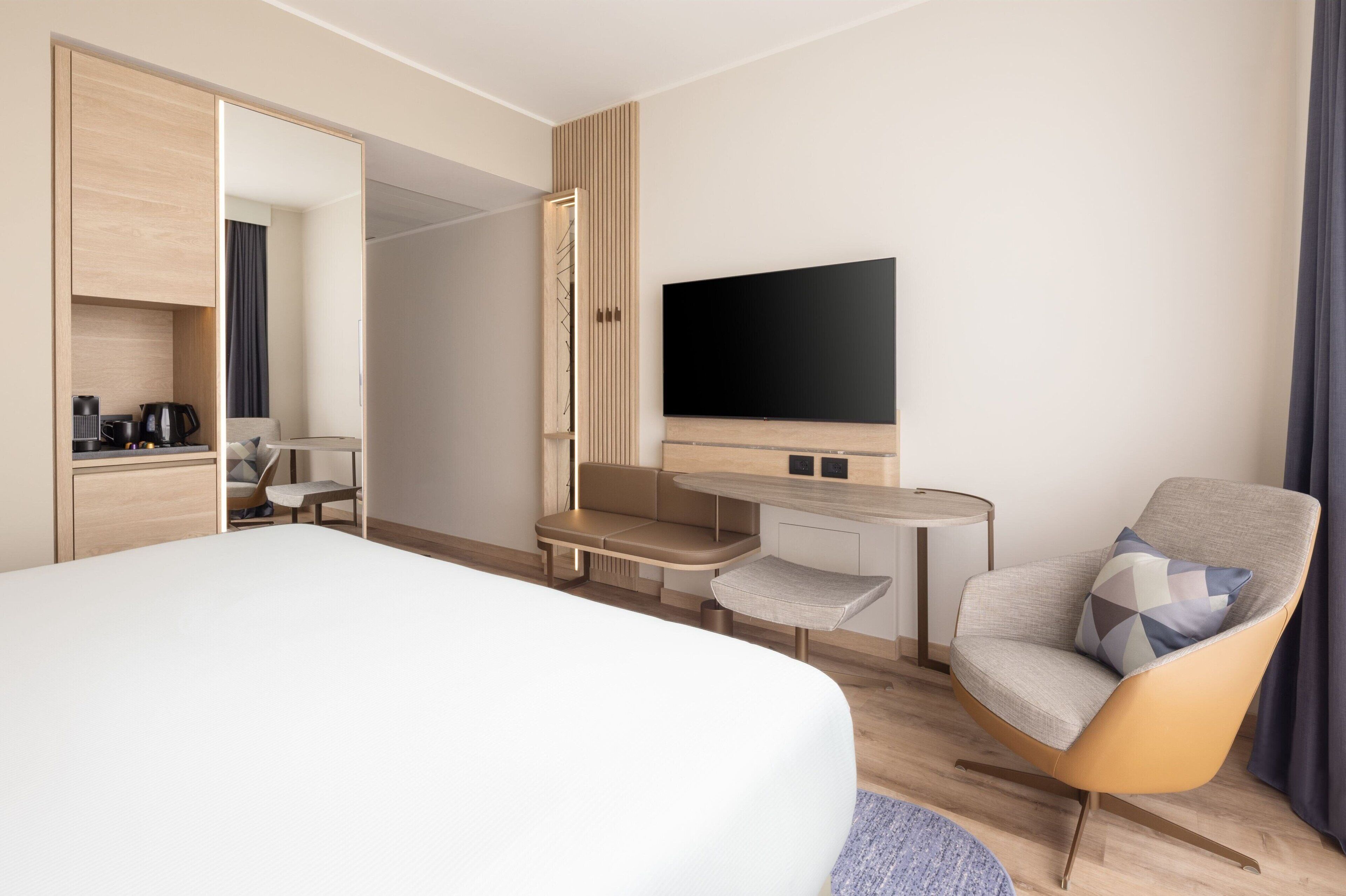 A modern hotel room with a bed, a desk, a chair, a TV, and a wardrobe.