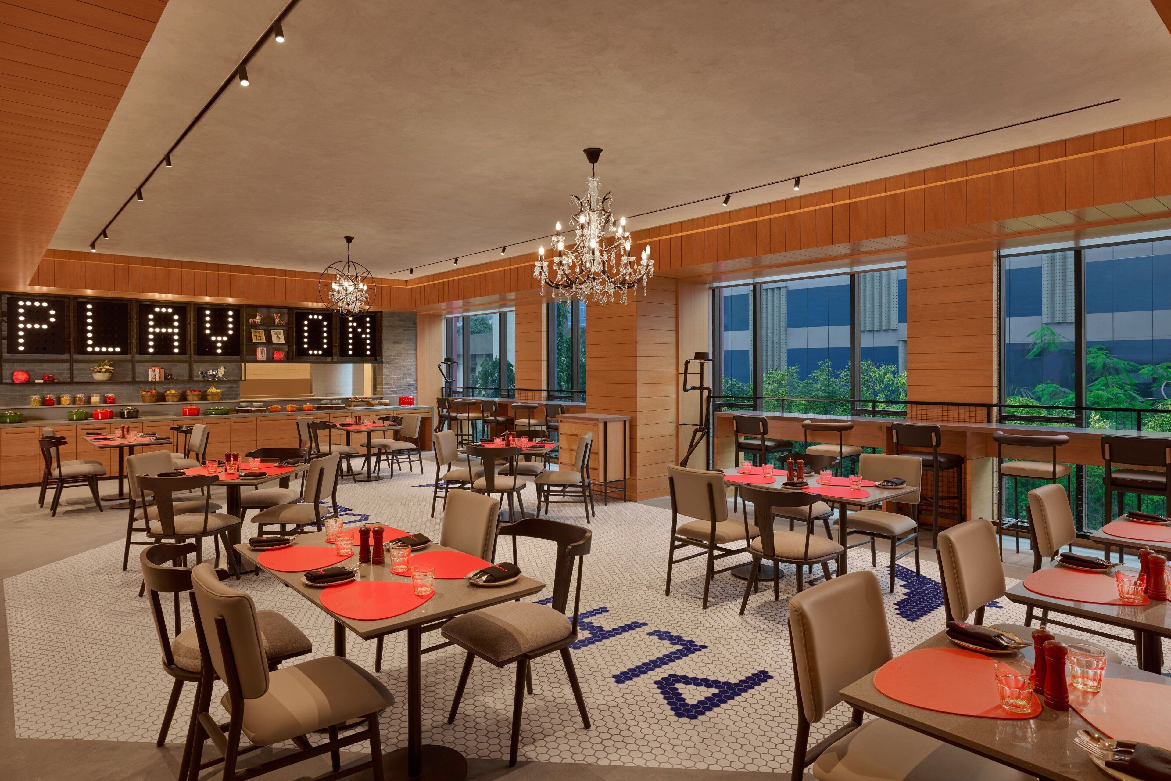 A spacious dining area with multiple tables and chairs, a bar area with a sign reading "PLAYON," and large windows offering a view of greenery outside.