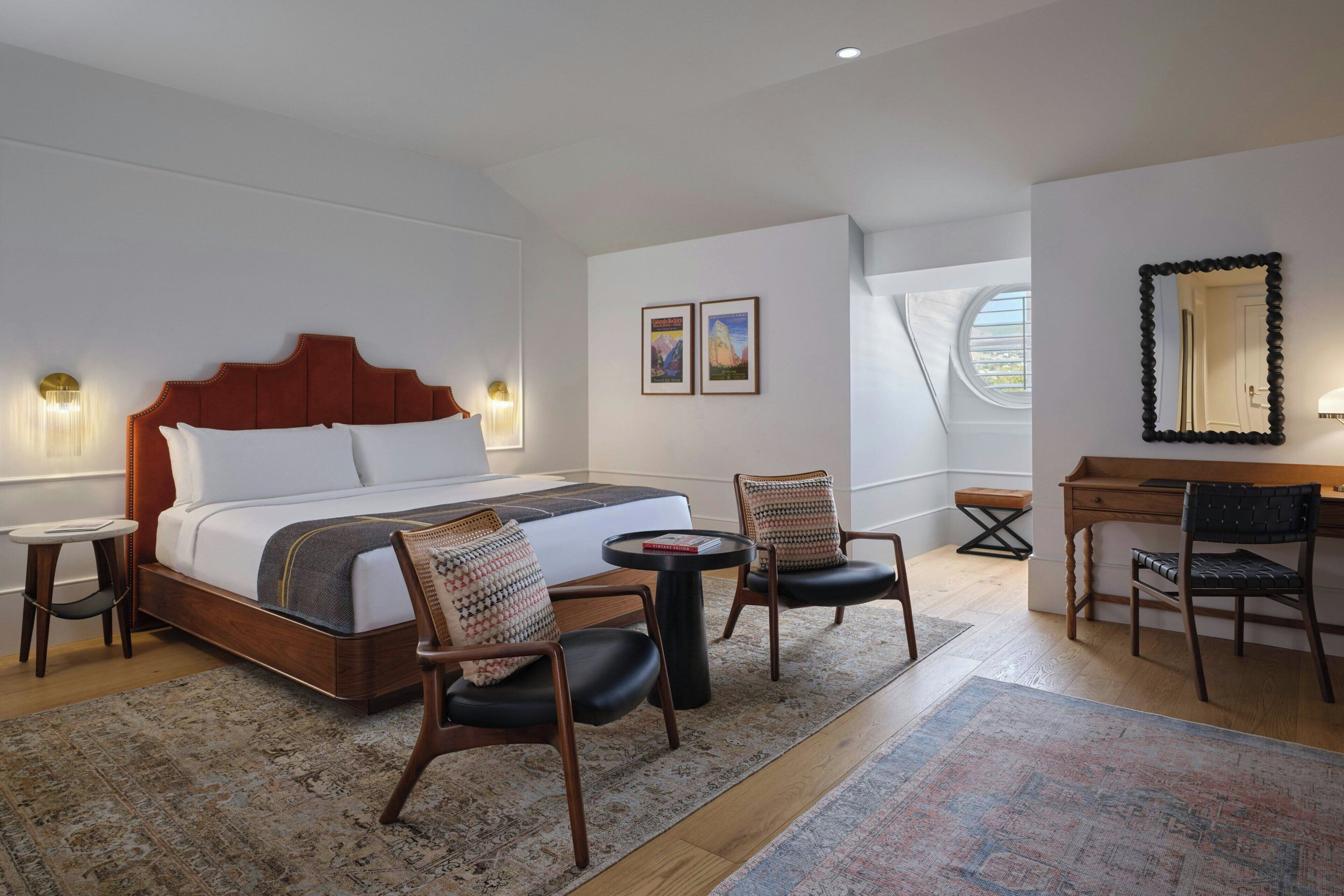 Quarto de hotel com cama, duas cadeiras, escrivaninha e janela.