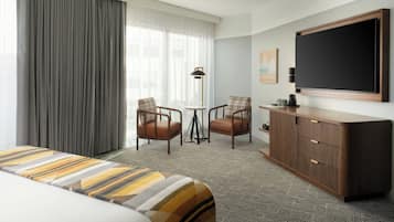 Quarto de hotel com uma cama, duas poltronas, uma mesinha, um armário e uma televisão.