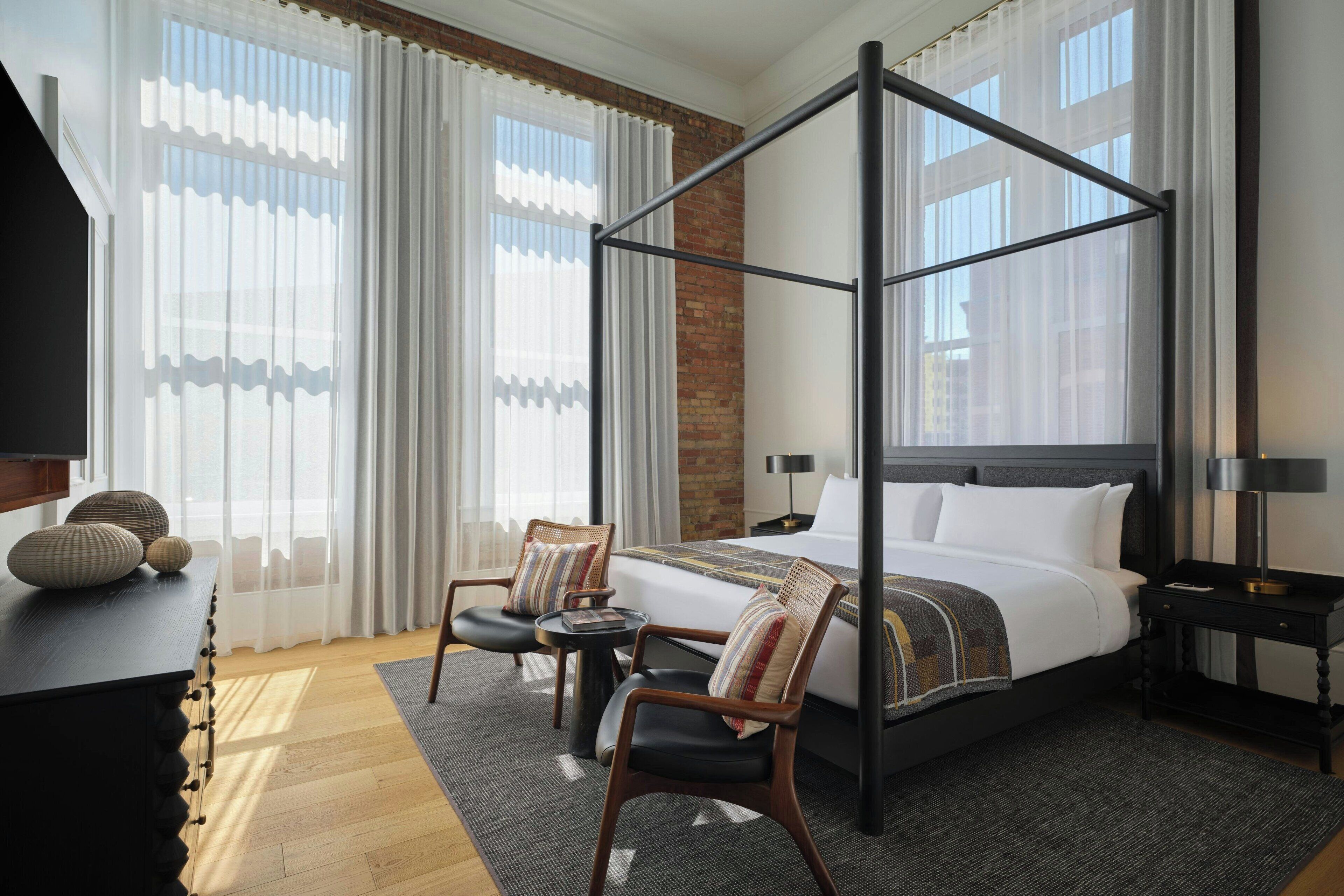 A hotel room with a four-poster bed, a desk with a chair, a small table, and a nightstand with a lamp.