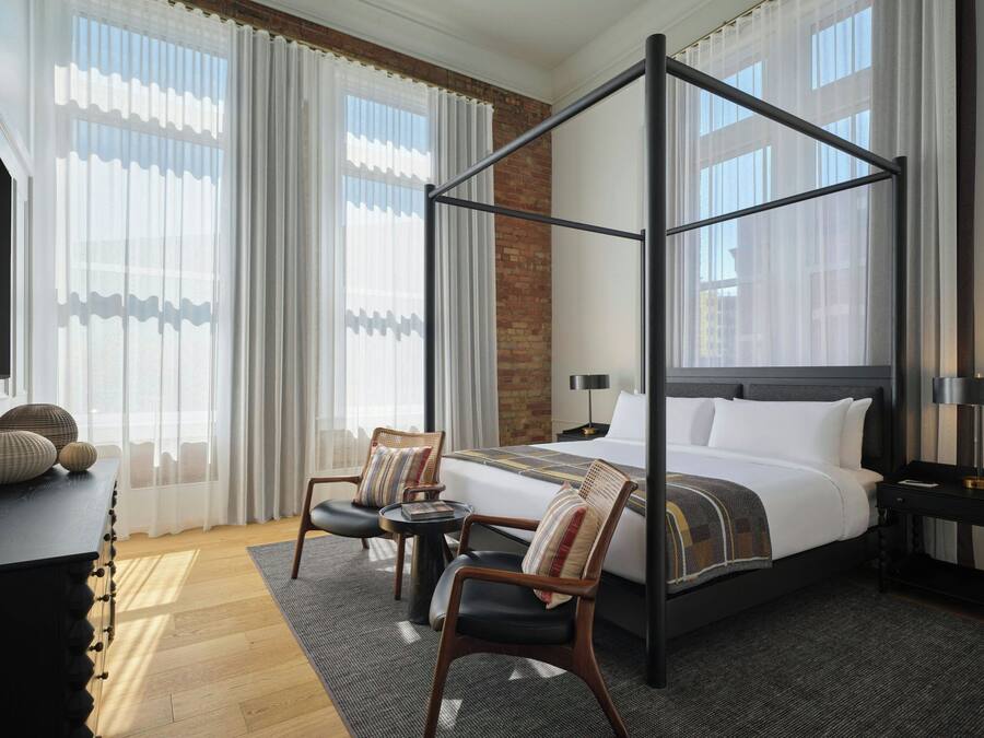 A hotel room with a four-poster bed, a desk with a chair, a small table, and a nightstand with a lamp.