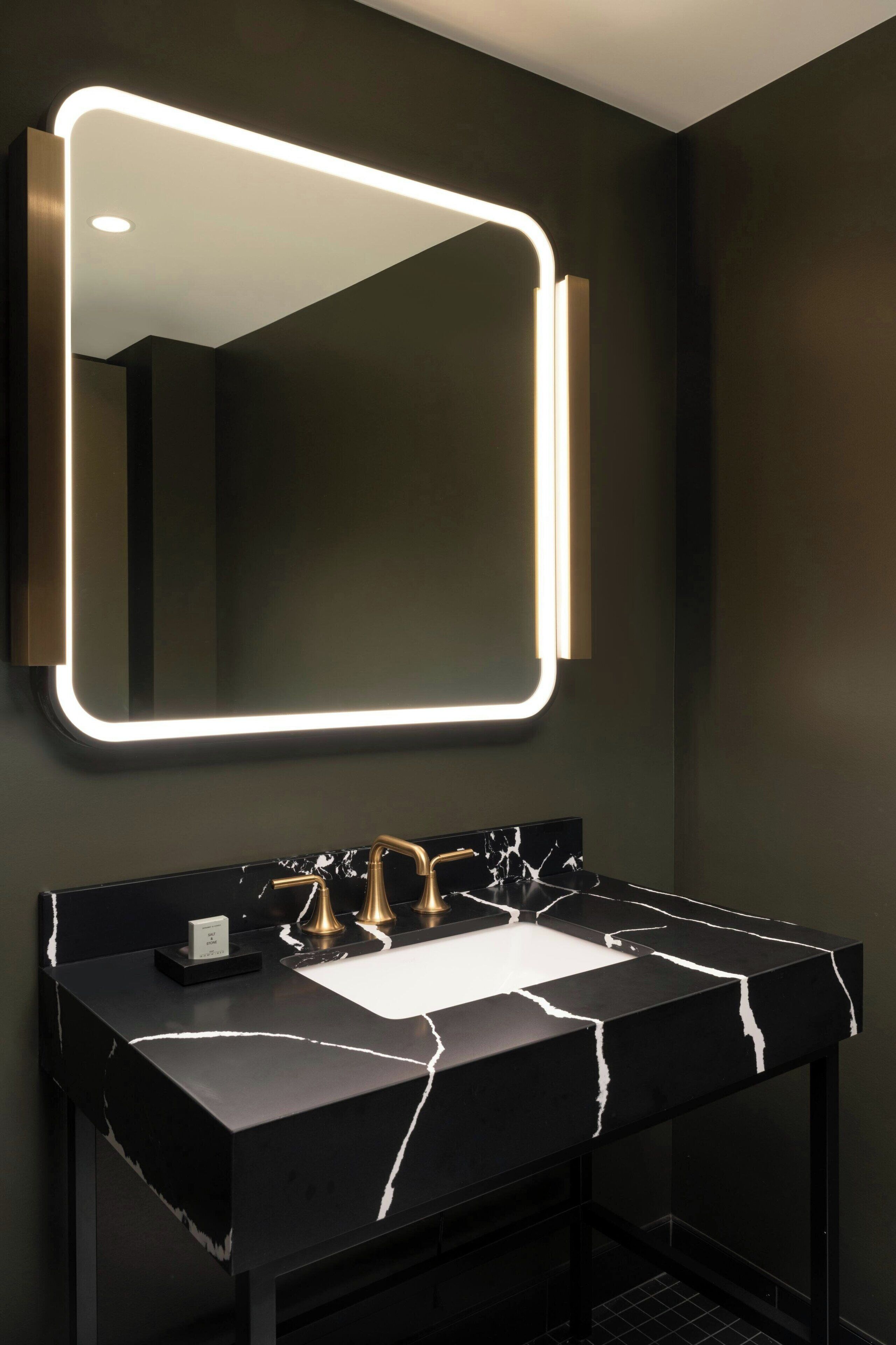 A modern bathroom with a black marble sink, gold faucets, and a wall-mounted mirror with illuminated edges.