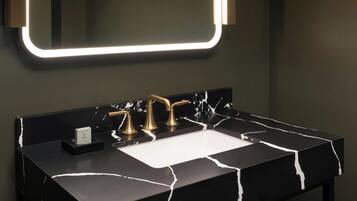 A modern bathroom with a black marble sink, gold faucets, and a wall-mounted mirror with illuminated edges.