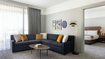 A modern hotel room with a dark-colored sofa, a round coffee table, and a large window with sheer curtains.