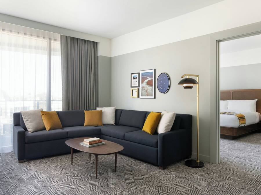 A modern hotel room with a dark-colored sofa, a round coffee table, and a large window with sheer curtains.
