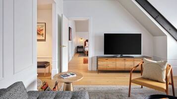 A modern living room with a television, a sofa, and a chair. There is a wooden cabinet and a small table with a book. The room has a high ceiling and a sloped ceiling with a wooden beam.