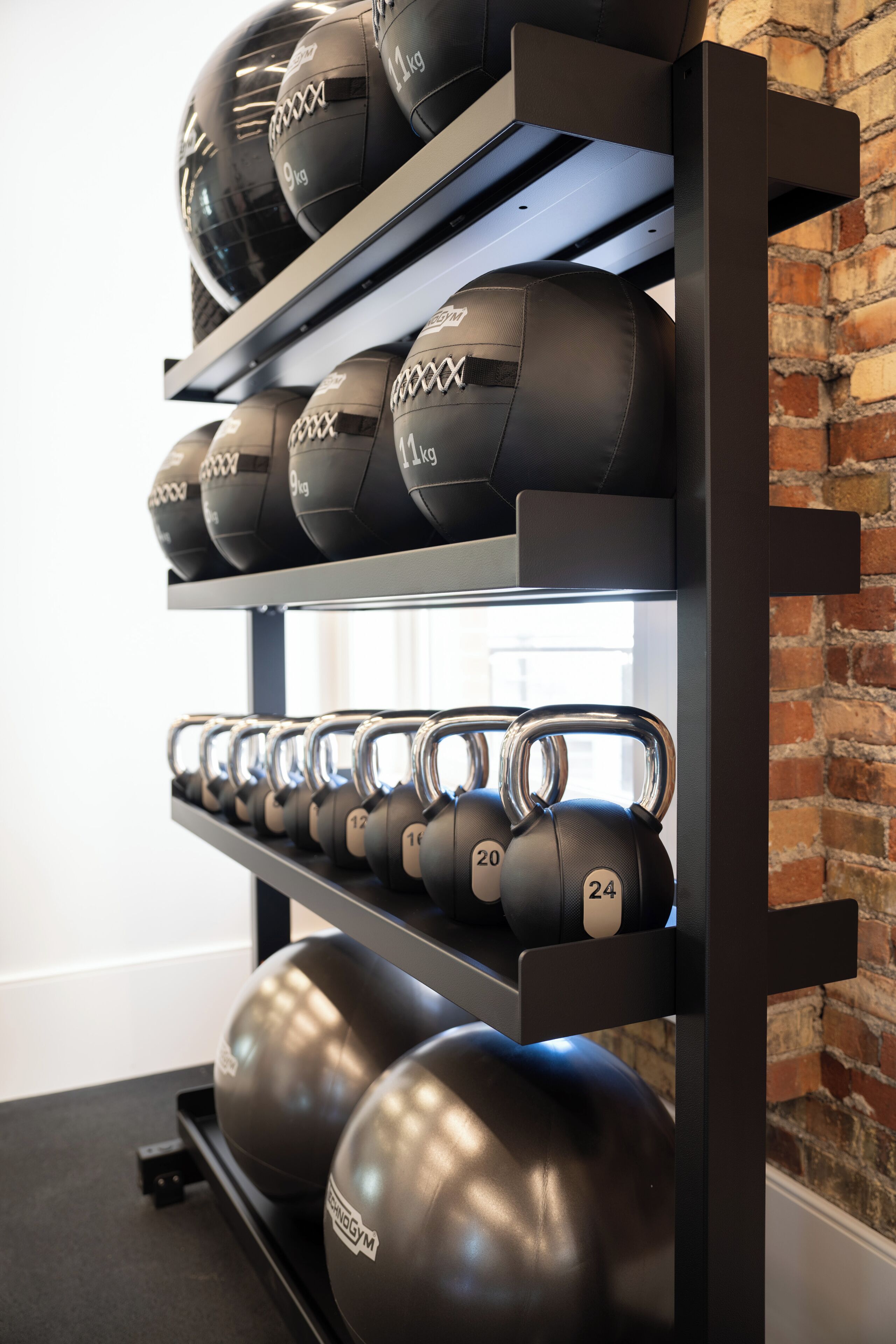 A wall-mounted rack with various weights, including kettlebells and dumbbells, against a brick wall.