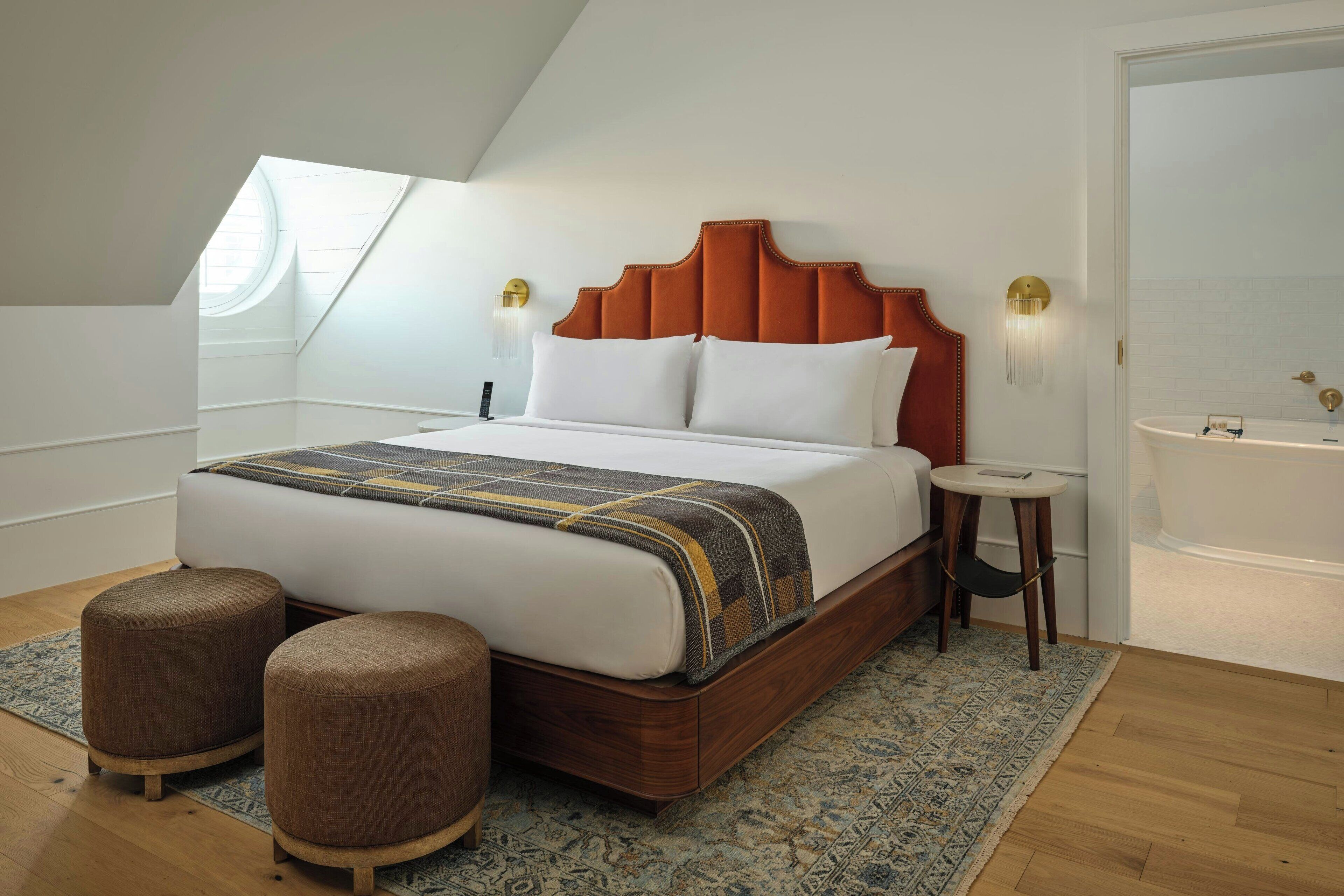 Een slaapkamer met een groot bed, twee voetenbankjes, een rond raam, een dakraam, een houten hoofdbord, een klein rond tafeltje en een badkamer met een bad.