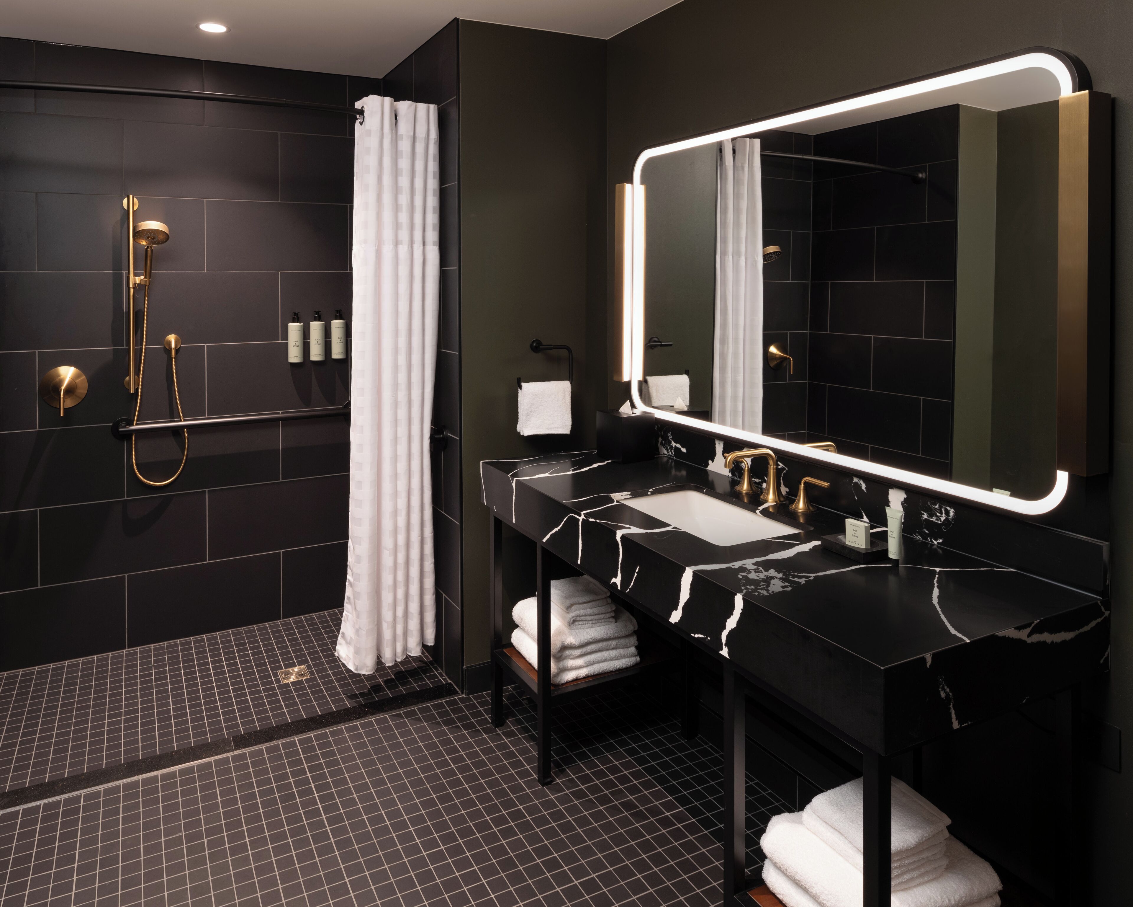 A modern bathroom with a black and white marble sink, a large mirror with integrated lighting, a gold showerhead, and a black tiled floor.