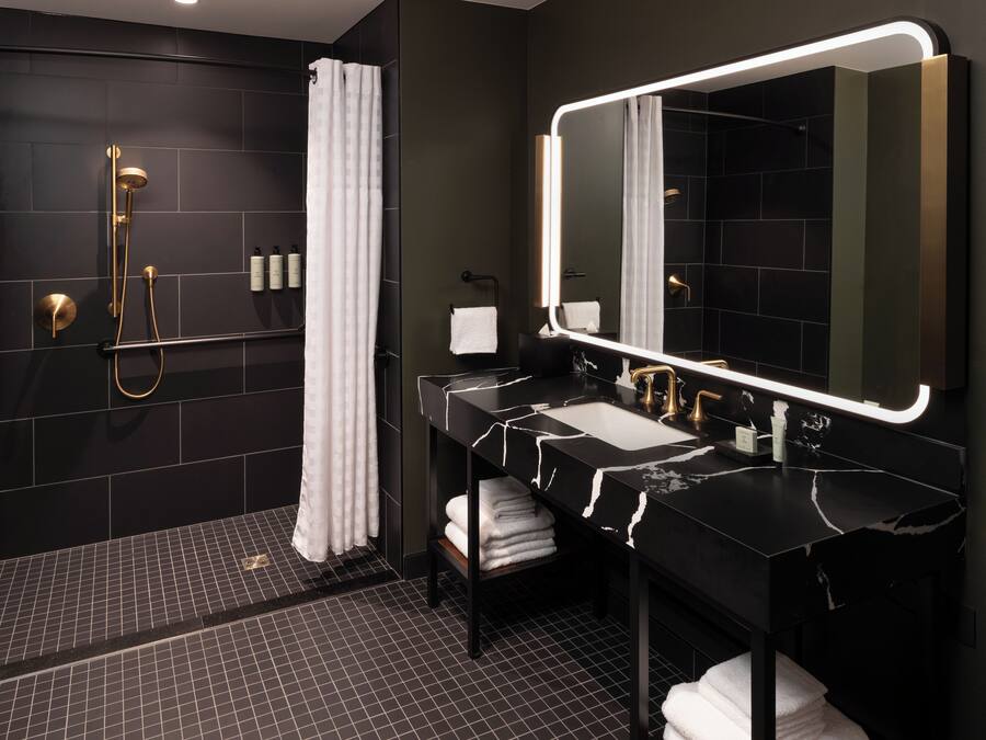 A modern bathroom with a black and white marble sink, a large mirror with integrated lighting, a gold showerhead, and a black tiled floor.
