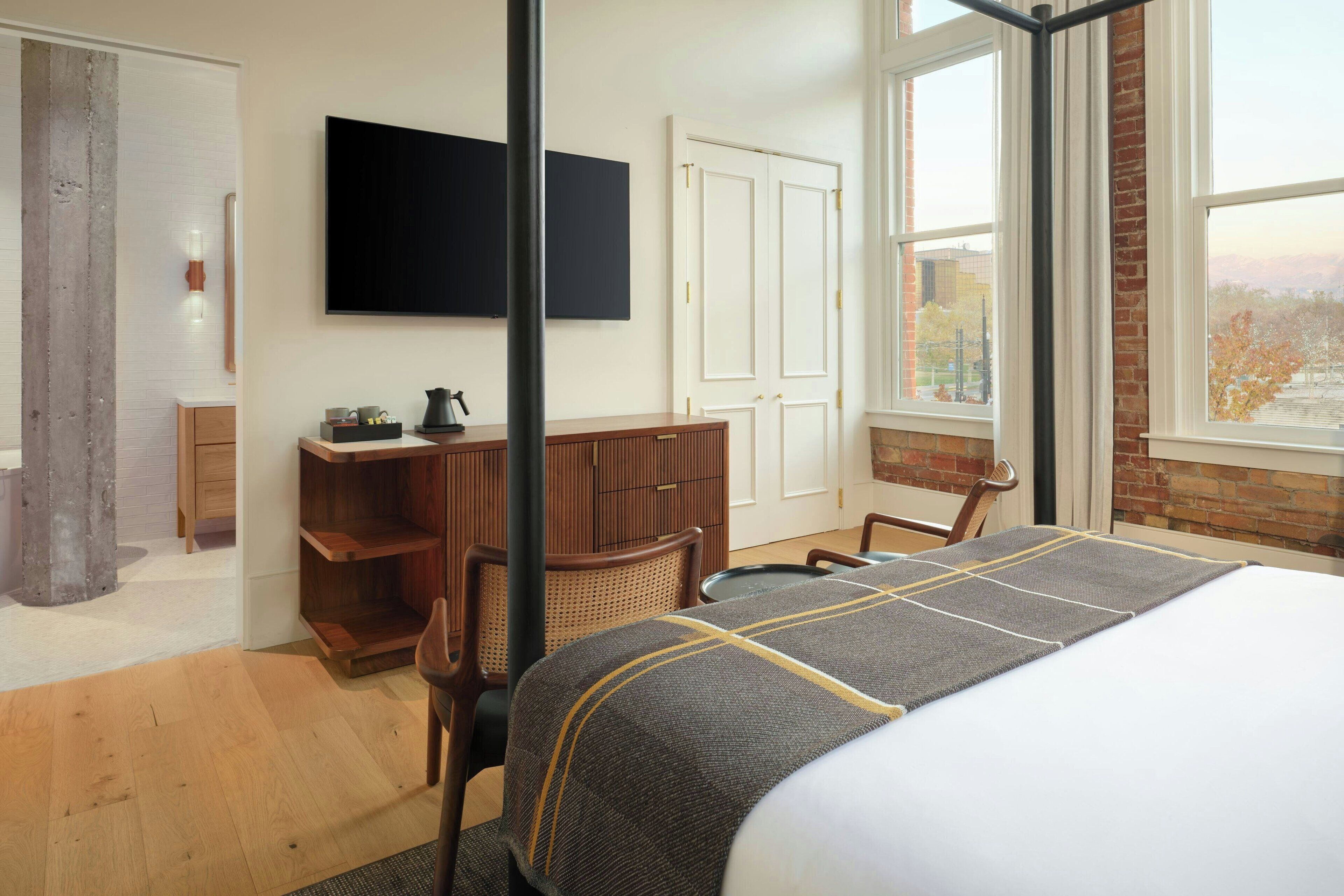 A hotel room with a bed, a desk, a chair, a television, and a window with a view of trees.