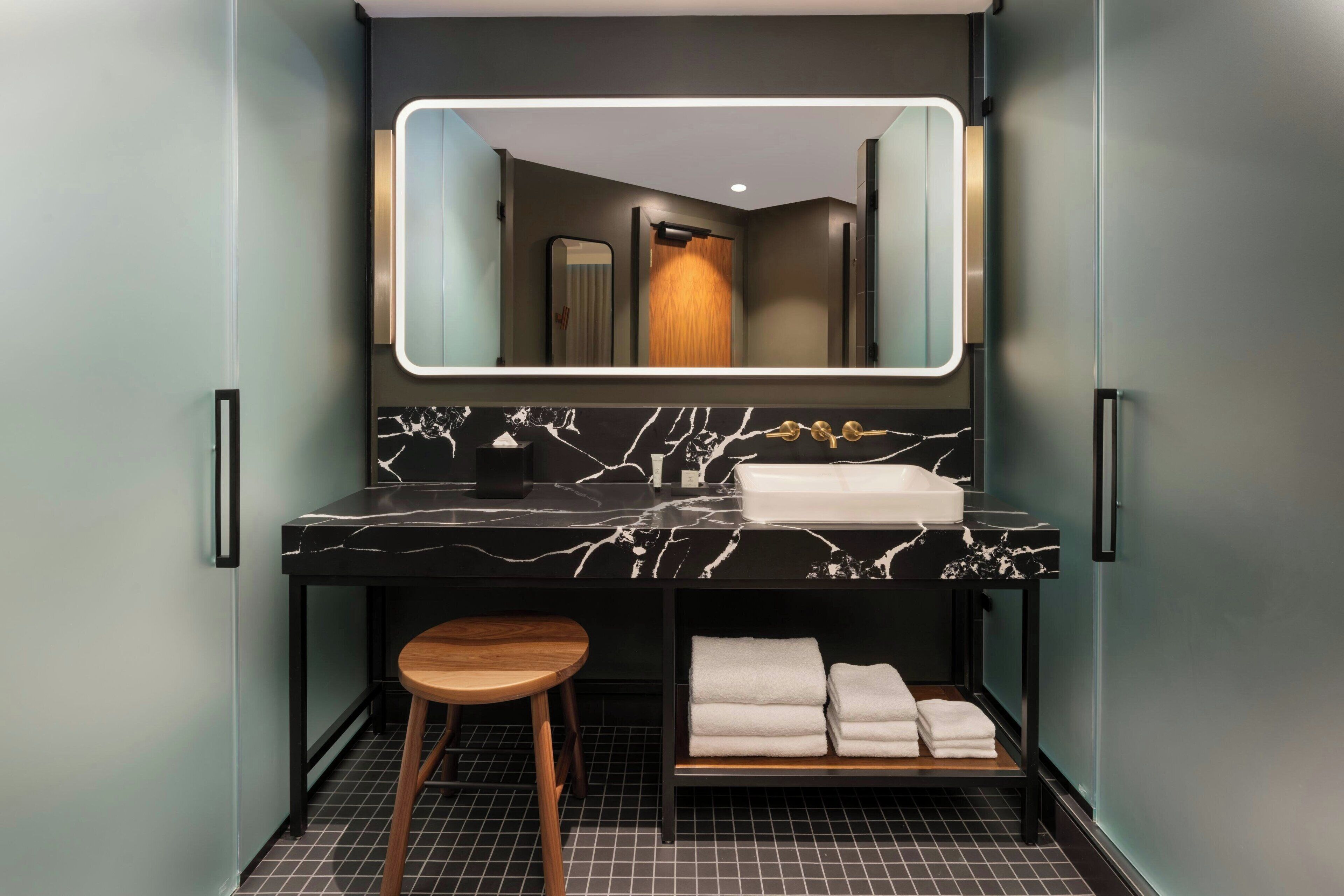 A modern bathroom with a black marble countertop, a large mirror, a wooden stool, and neatly folded towels.