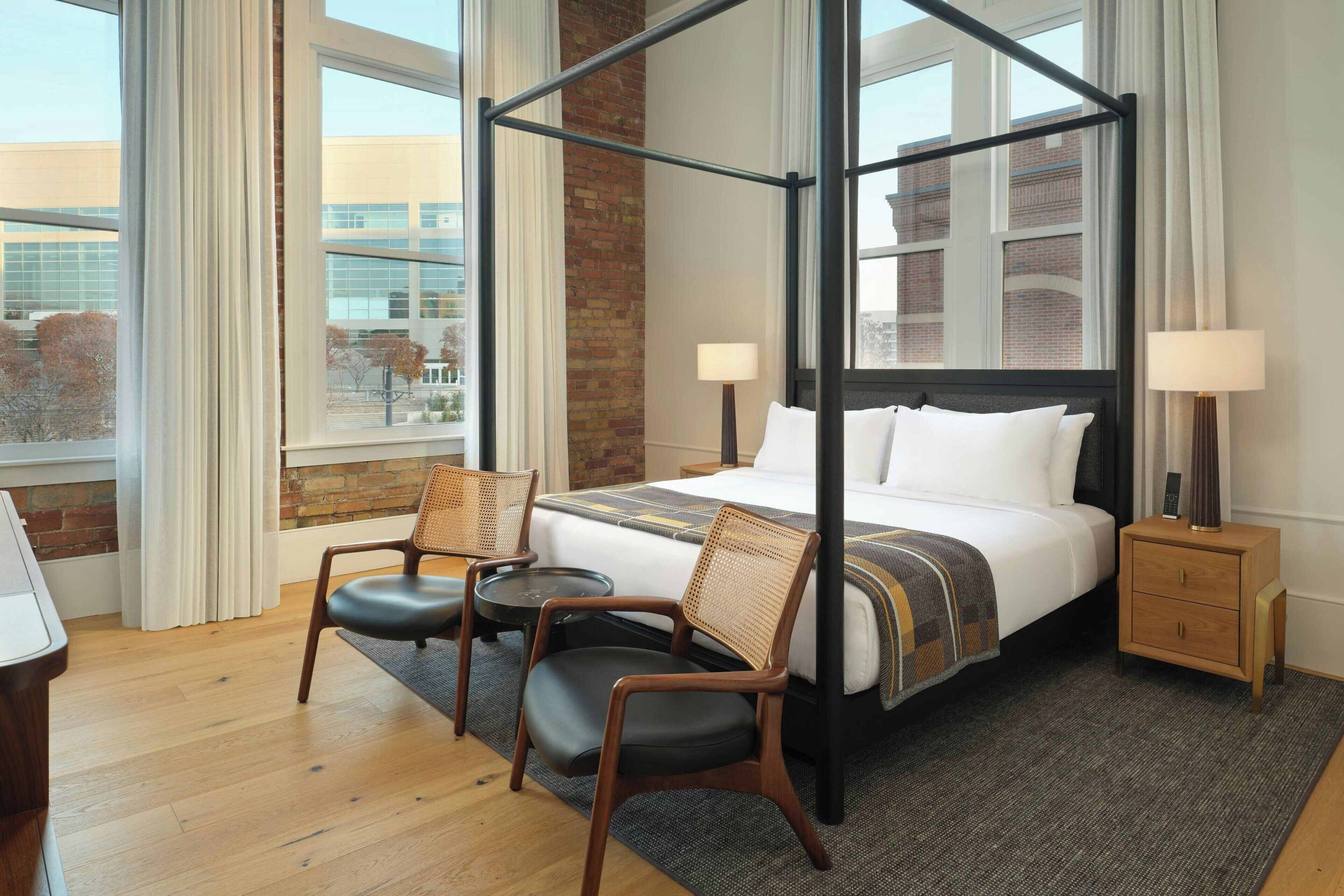 A four-poster bed with a wooden frame, a small bedside table with a lamp, two wicker chairs, and a view of a brick building through large windows.