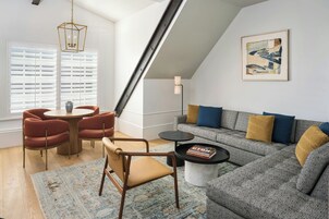 Una sala moderna con techo estilo loft, un sofá gris con cojines azules y mostaza, una mesa de centro redonda, y un comedor con sillas rojas y una mesa de madera.