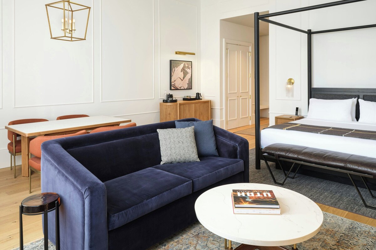 A modern hotel room with a four-poster bed, a dark blue sofa, a round coffee table, and a dining area with a wooden table and chairs.