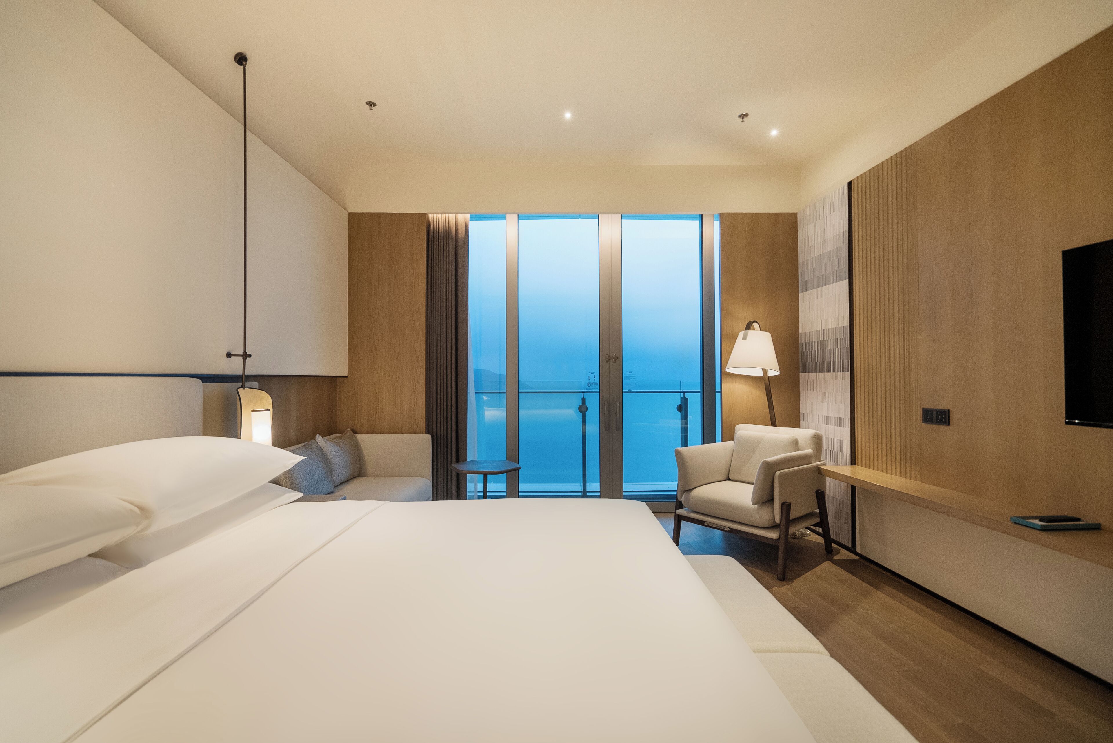 Una habitación de hotel moderna con una cama grande, un escritorio, una silla y vistas al mar a través de ventanales de gran altura.