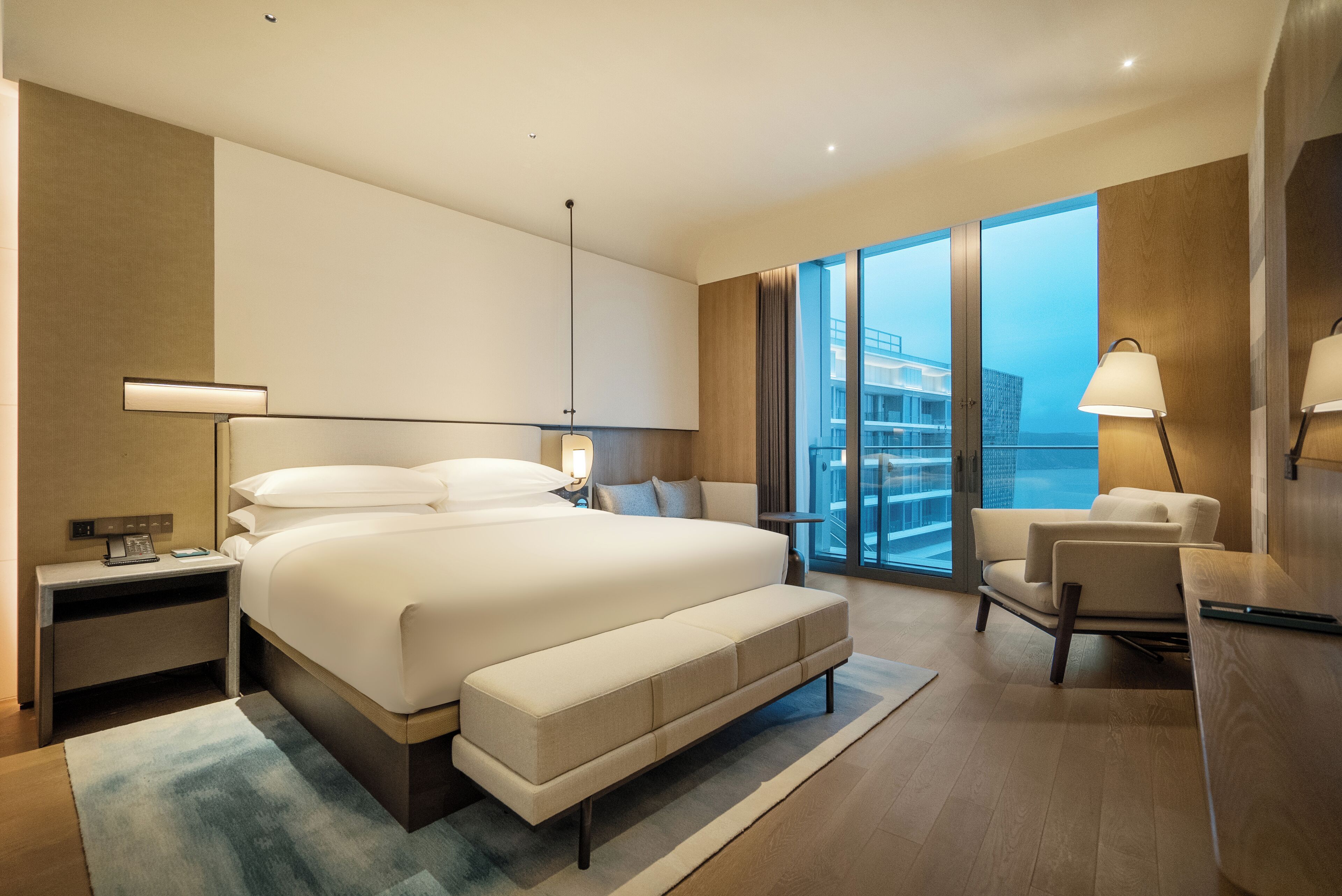 Une chambre d’hôtel moderne dotée d’un grand lit, d’un coin salon, d’un bureau et offrant une vue sur la ville grâce à de grandes fenêtres.