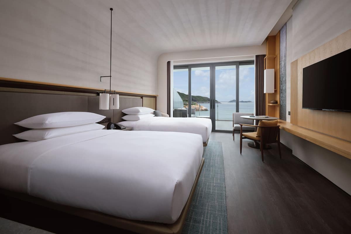 A hotel room with two beds, a desk, a chair, a TV, and a large window offering a view of the sea.