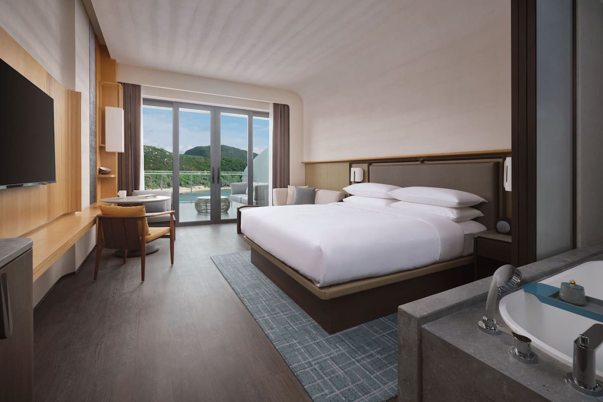 A modern hotel room with a large bed, a desk with a chair, a TV, a bathroom with a bathtub, and a balcony with a view of the hills.