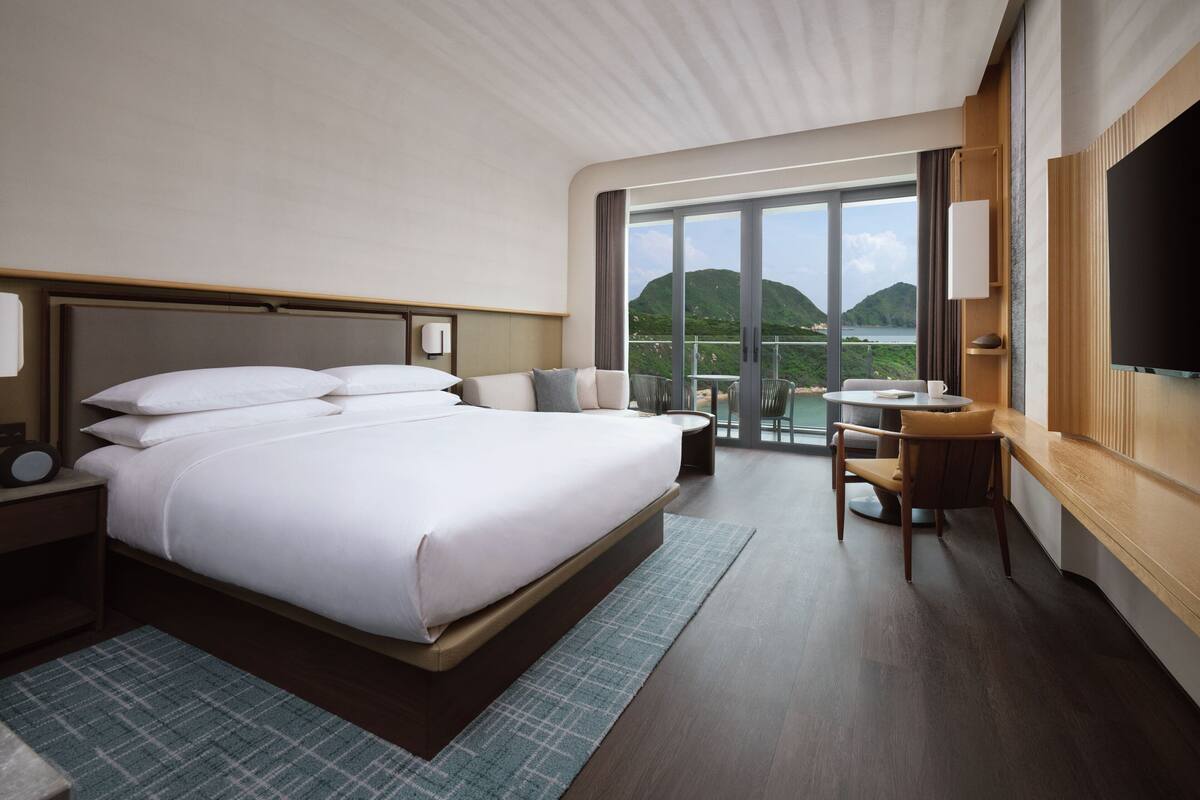 A modern hotel room with a large bed, a desk with a chair, a TV, and a balcony with a view of hills.