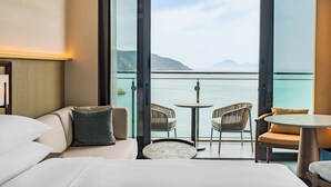 Habitación de hotel con un amplio ventanal corredera que da a un balcón con vistas al mar y a las montañas. Incluye una cama, una mesita redonda y dos sillones de mimbre.