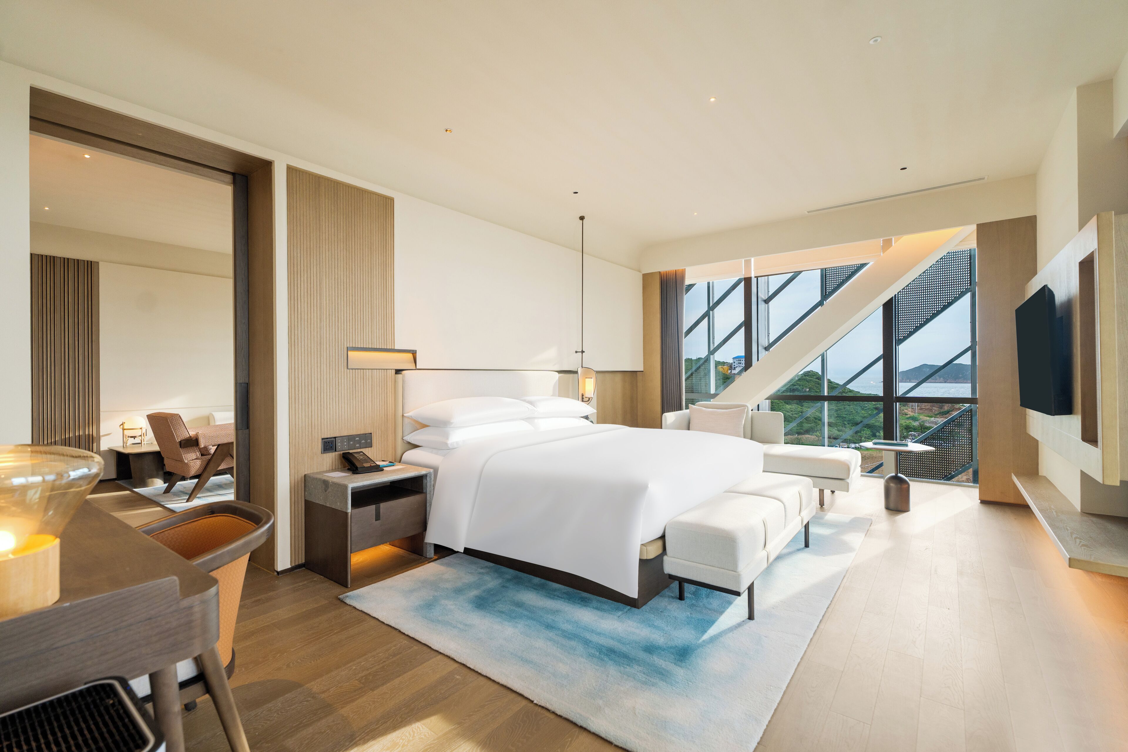 A modern hotel room with a large bed, a desk with a chair, a TV, and a view of the outdoors.