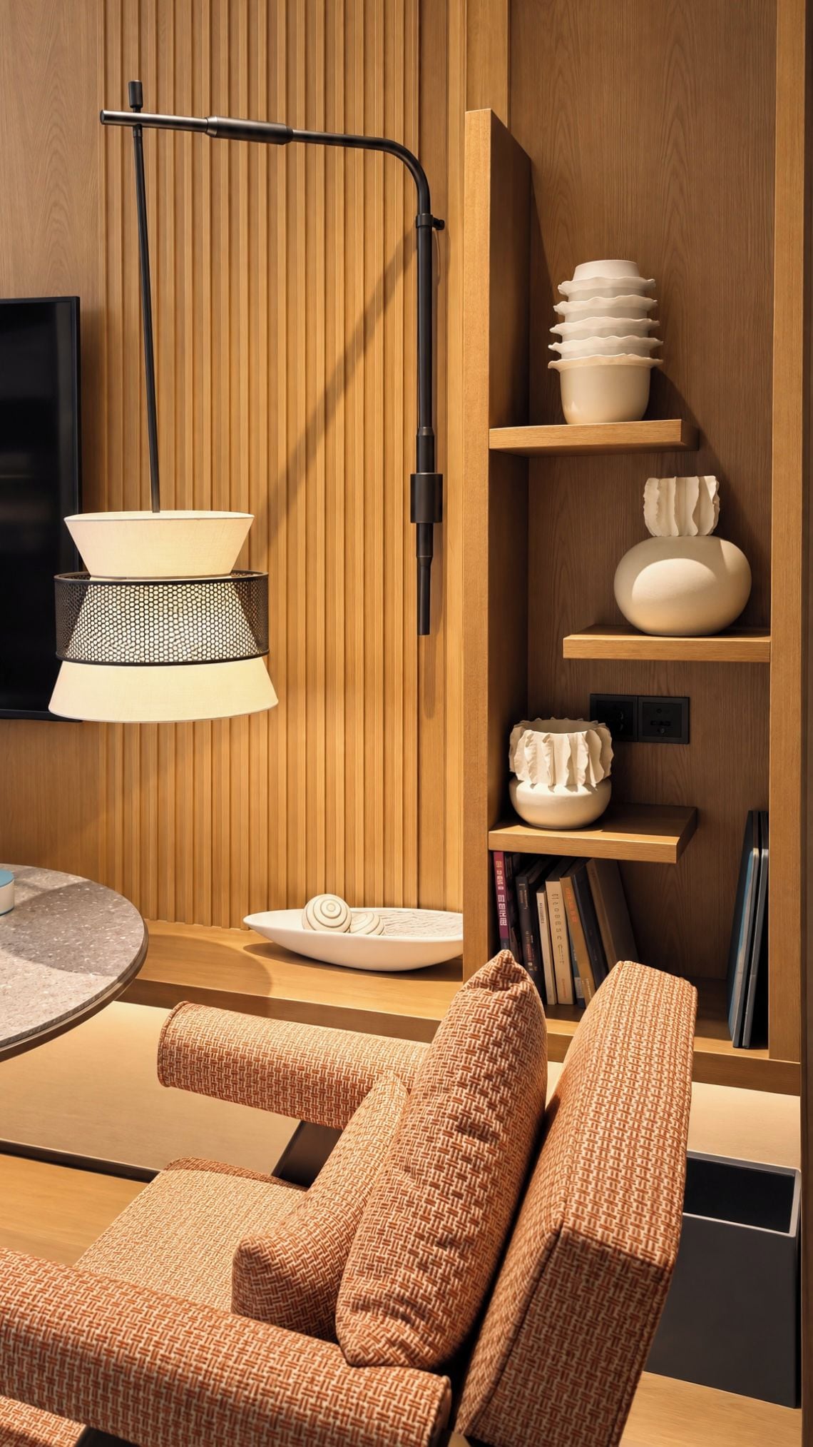 A modern interior with a round table, two patterned armchairs, a wall-mounted lamp, and wooden shelves with decorative items.