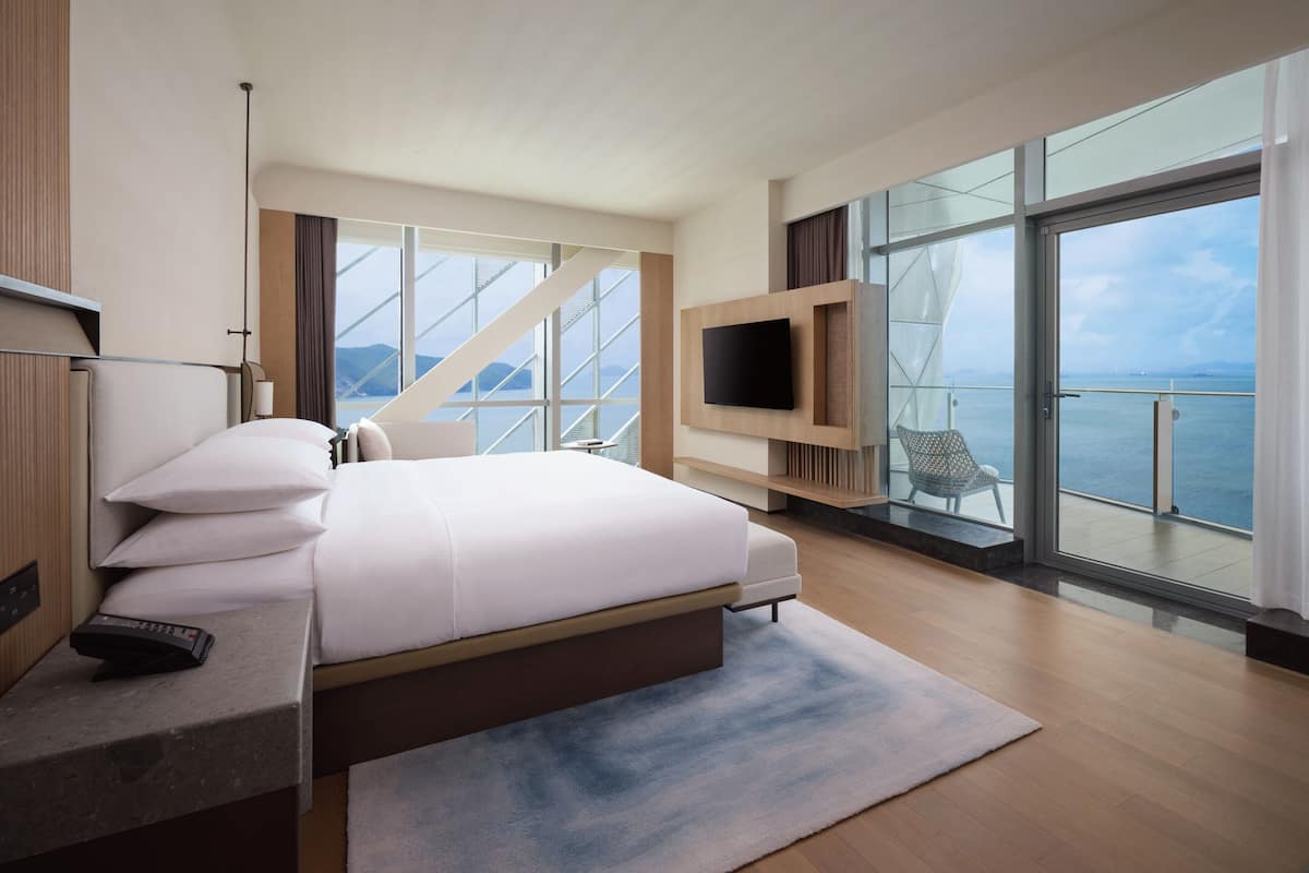 A modern hotel room with a large bed, a TV mounted on the wall, and a balcony with a view of the sea.