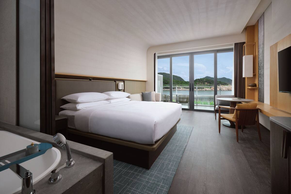 A modern hotel room with a large bed, a bathroom with a bathtub, a desk, and a view of the sea.