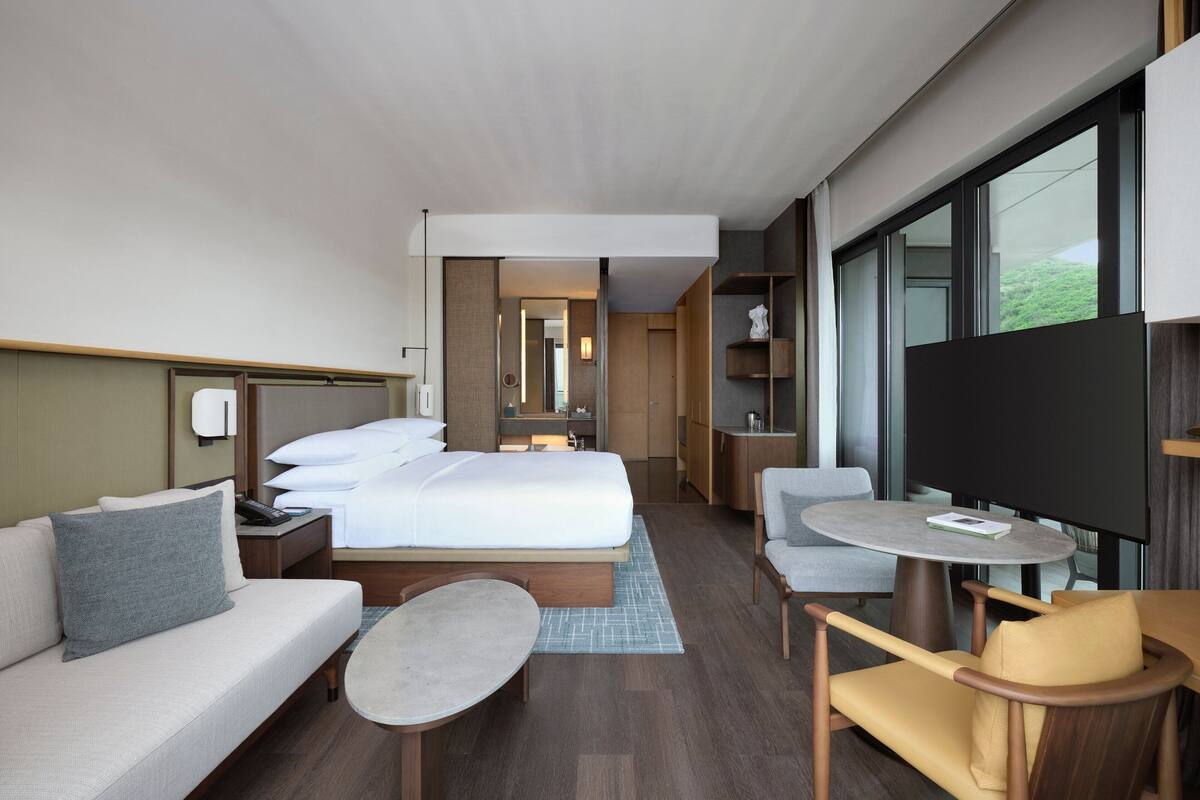 A modern hotel room with a bed, sofa, armchair, and a small table. There is a TV and a large window providing a view of the outdoors.
