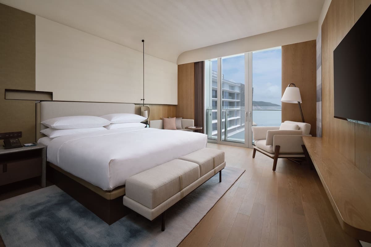 A modern hotel room with a large bed, a seating area, a TV, and a balcony with a view.