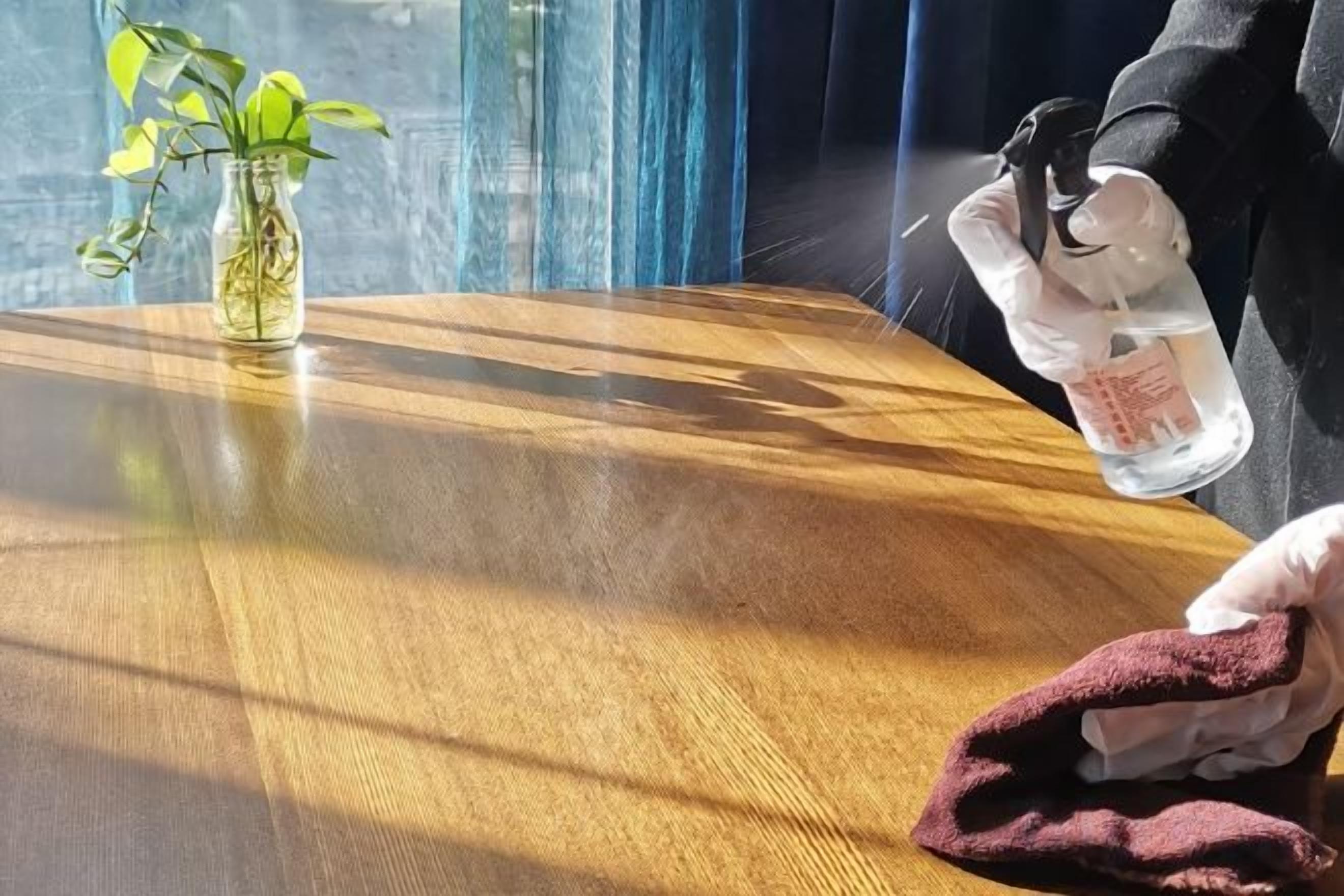 Une table en bois sur laquelle se trouve un vase en verre contenant une plante, une personne tenant un verre d’eau et un tissu.