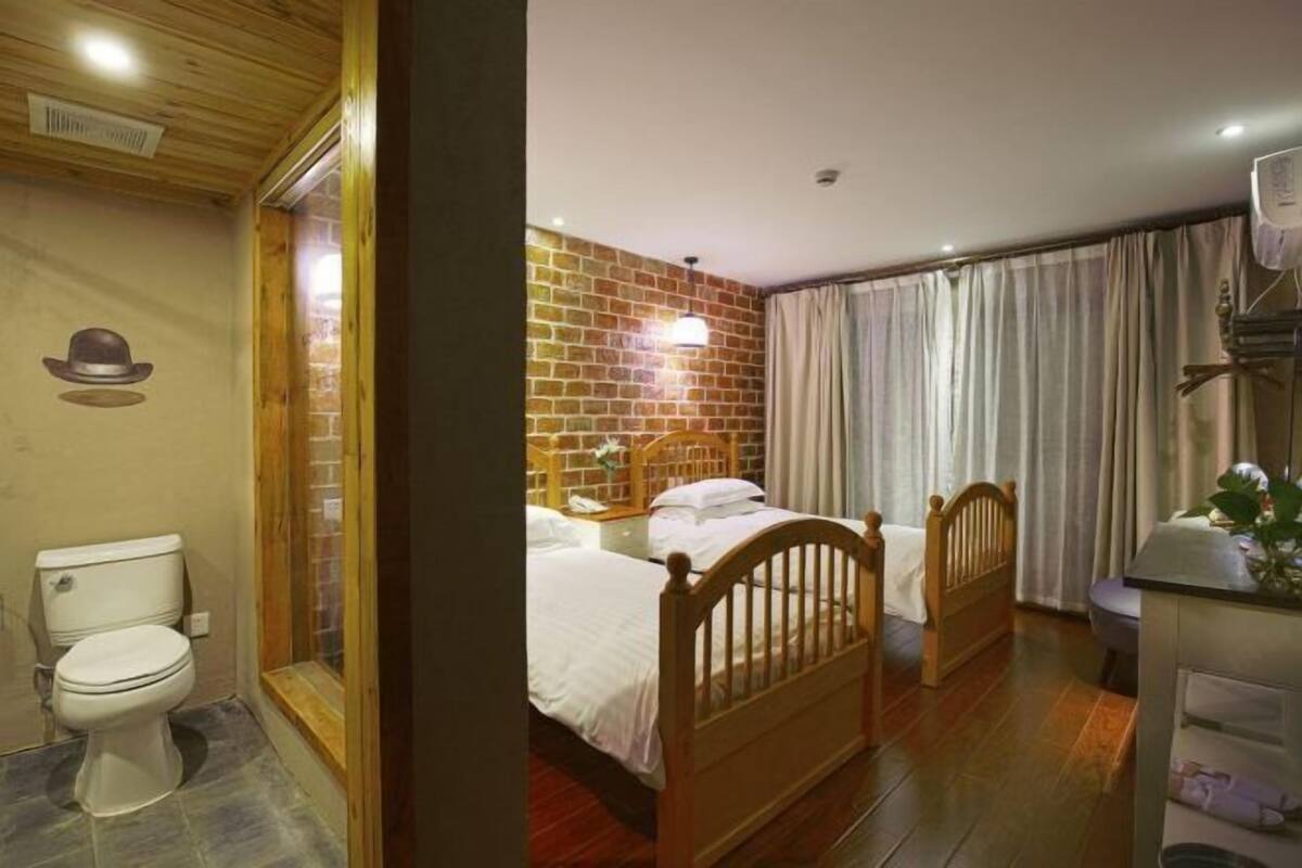 Una stanza con soffitto in legno, parete in mattoni, due letti con testiere in legno, un WC bianco e una finestra con tende.