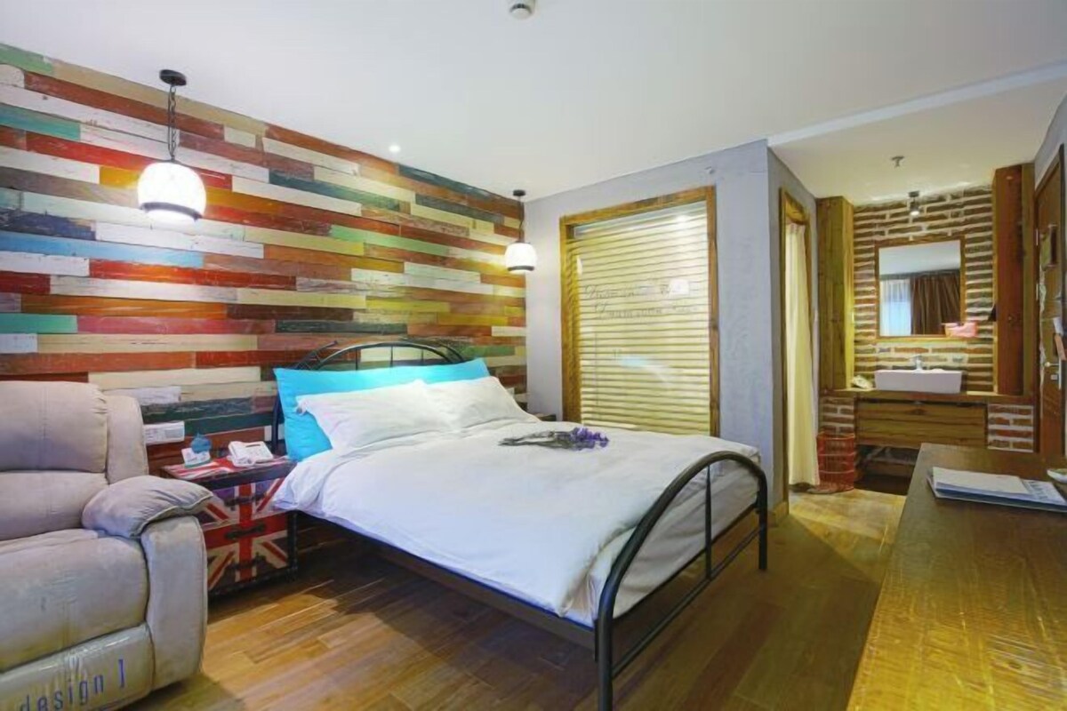 A bedroom with a bed, a chair, a wooden wall with colorful planks, a window with blinds, and a bathroom with a mirror and sink.