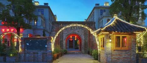 A traditional building with a stone archway entrance, a small adjacent structure with a tiled roof, and a paved walkway leading to the entrance.