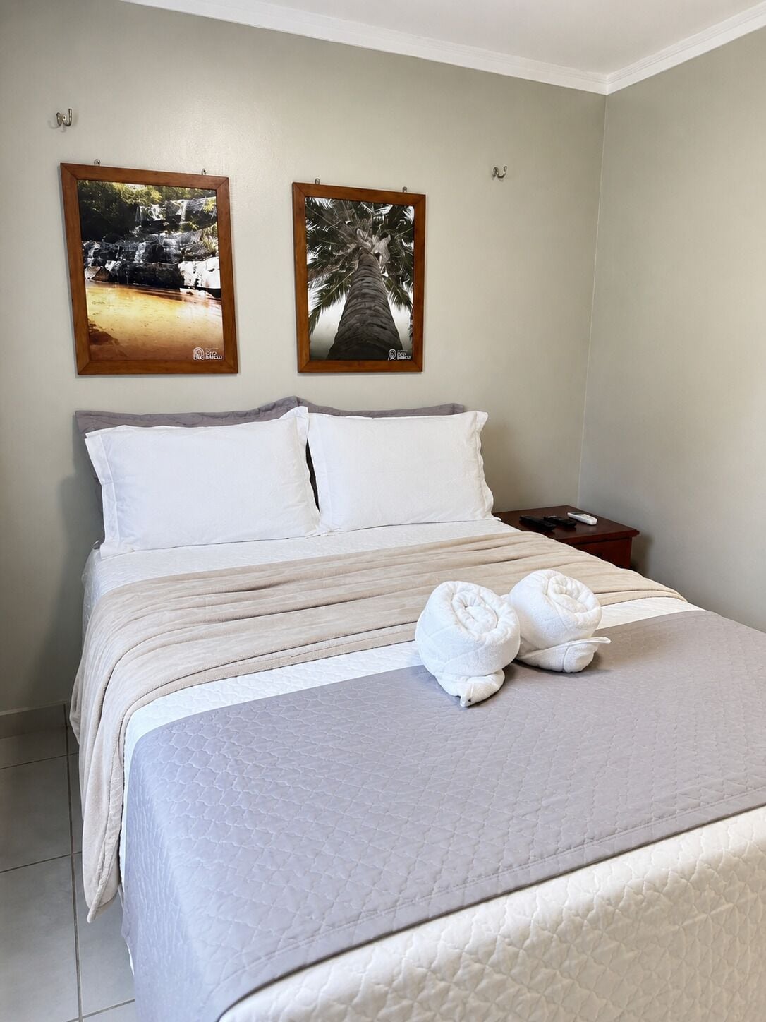A neatly made bed with white linens, two wall-mounted pictures, and a small bedside table with a remote control.