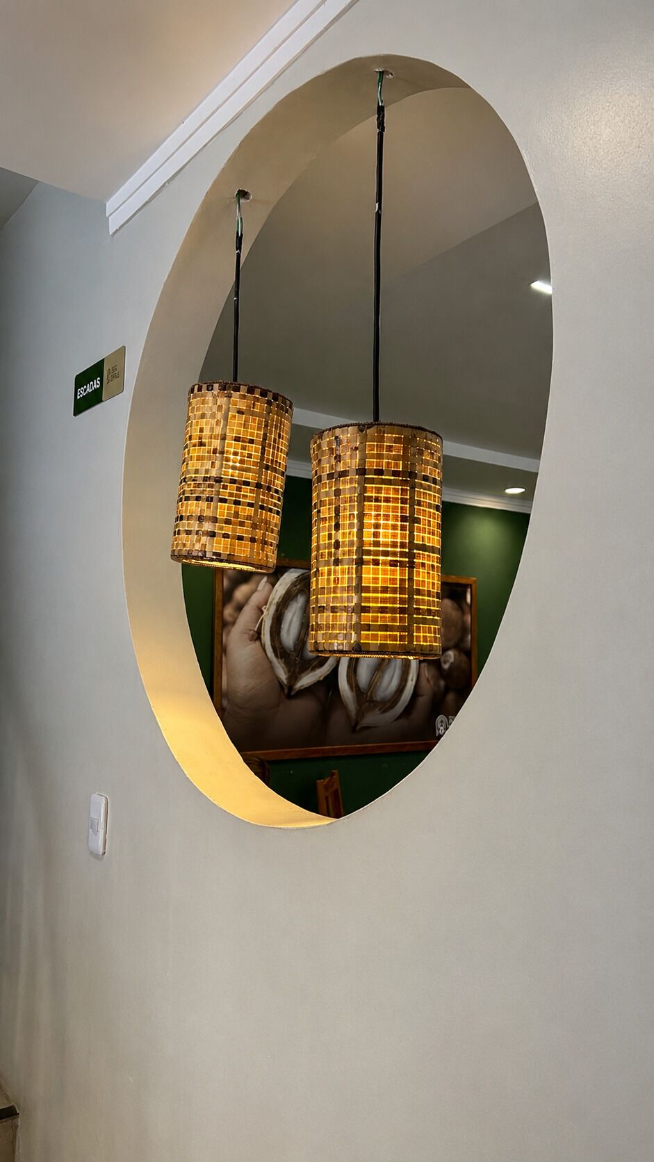 A circular mirror with two hanging lamps, a sign indicating 'Escadas,' and a partial view of a room with a brown leather sofa.