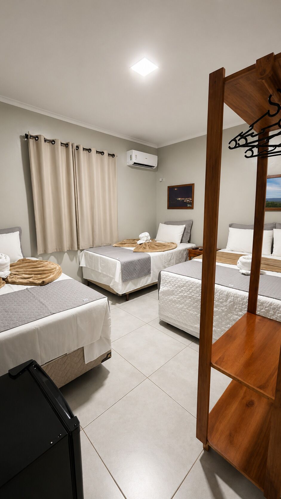 A hotel room with three beds, a wooden bench, a black suitcase, and a wall-mounted air conditioner.