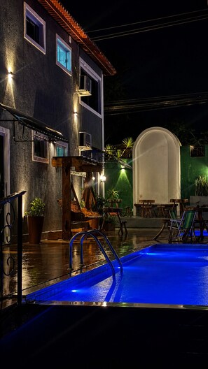 A pool area with a blue-lit pool, a wooden deck, and a building with a tiled roof. - Pousada Coco Babaçu (Vicosa do Ceara)