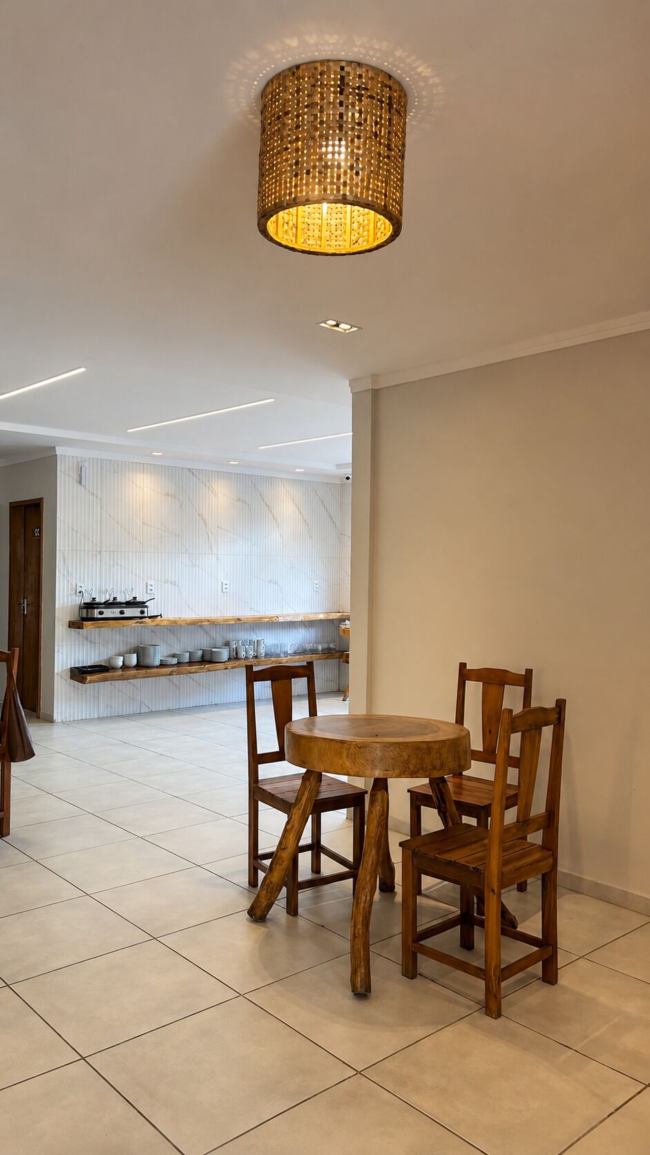 A dining area with a round wooden table and chairs, a woven pendant light, and a kitchen in the background.